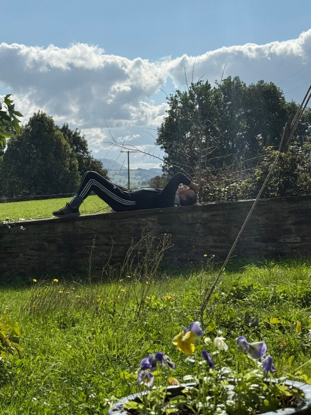 Man relaxes in sunshine lying on a low all amidst fields, trees and a sunny sky with clouds