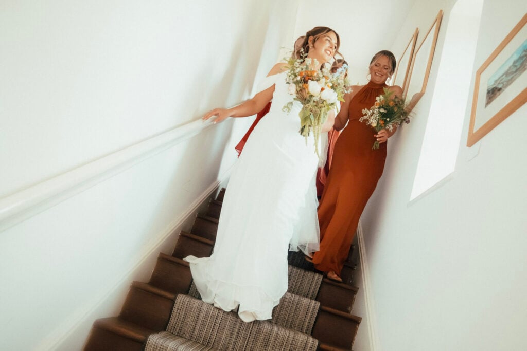 Bride with bouquet and bridesmaids wearing burnt orange gowns walking down stairs in The Collquy at White Heron Estate wedding venue Herefordshire
