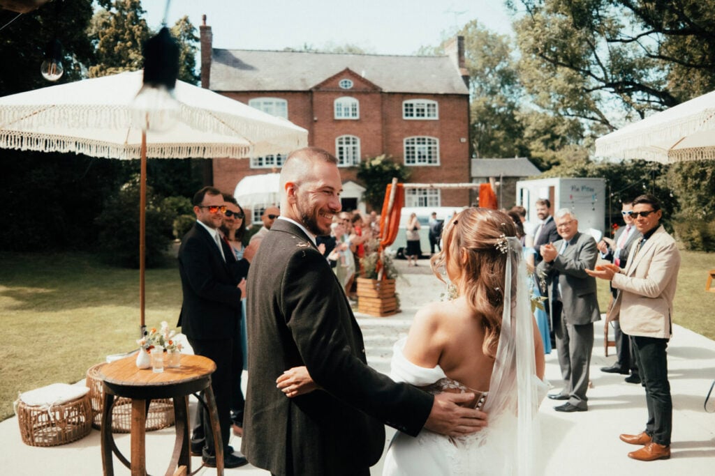 A Bride and Groom smiling in front of their guests outside The Sheriffs at White Heron Estate in Herefordshire
